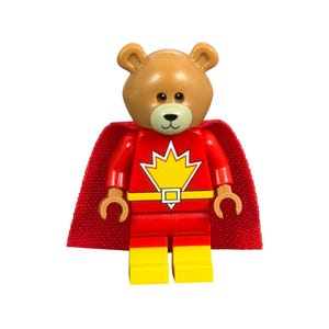 May include: A brown bear figure with a red superhero suit, yellow accents, and a red cape. The bear has a black nose and eyes, and the suit features a yellow emblem. The figure is a toy.