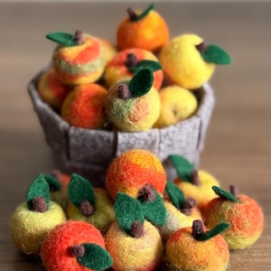 May include: A collection of handmade felt apples in various colors, including red, yellow, and orange, with green felt leaves and brown stems. Some apples are in a gray basket, while others are scattered on a wooden surface.
