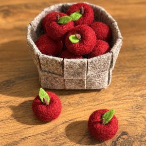 May include: A woven light brown felt basket filled with several small, round, red felt apples, each featuring a tiny brown stem and a single green felt leaf. Two additional red felt apples with stems and green leaves are placed in front of the basket.