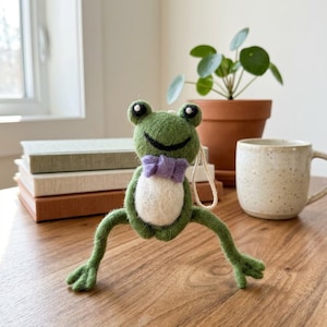 May include: A handmade green felt frog ornament with a white belly and a purple bow tie. The frog has large, round eyes and outstretched legs. A string is attached for hanging. The ornament is on a wooden surface.