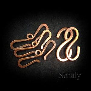 May include: Five copper S-shaped hooks, with two larger hooks in the foreground and three smaller hooks in the background. The hooks are arranged in a semi-circle shape. The background is black.