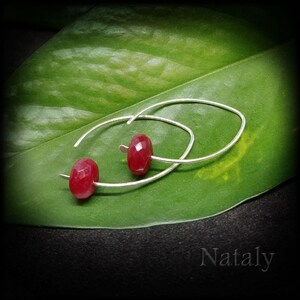 May include: A pair of silver hoop earrings with a red gemstone accent. The earrings are on a green leaf.