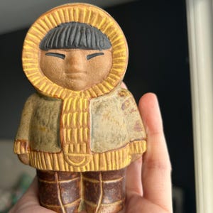 May include: A ceramic figurine of a person in a hooded parka. The figure has a serene expression, black hair, and is dressed in a yellow and brown outfit. The figurine is held in a hand.