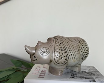 Op de afbeelding: Een keramieken neushoornbeeldje in grijze en beige tinten. De neushoorn heeft gedetailleerde kenmerken en staat op een stapel boeken. De boektitels omvatten "ELENA FERRANTE". Een decoratief item.