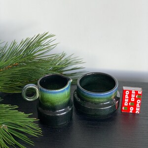 May include: A small ceramic creamer and sugar bowl set. The pieces are dark green with a blue and green gradient around the rim. The creamer has a handle. A red and white matchbox is next to the sugar bowl.