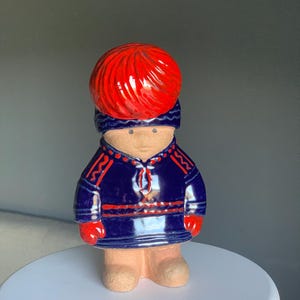 Peut inclure: Figurine en céramique représentant une personne vêtue d'un manteau bleu foncé avec des bordures rouges et des gants rouges. La figurine a une grosse coiffe rouge en forme de boule et des jambes et des pieds de couleur beige.