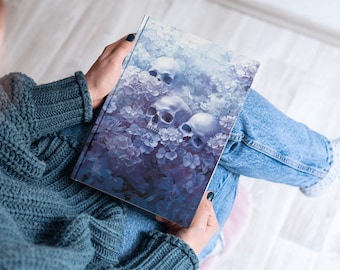 May include: A blue and purple notebook with a floral design and three white skulls. The notebook is being held by a person.