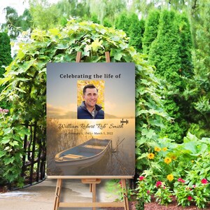 May include: A memorial sign with a photo of a man and the text "Celebrating the life of William Robert Rob" Smith February 5, 1945-March 5, 2022". The sign has a background image of a lake with a boat and a sunset.