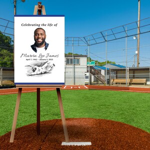 May include: A memorial sign on a wooden easel at a baseball field. The sign reads "Celebrating the life of Marvin Lee James, April 7, 1962 - January 2, 2023" with a portrait and baseball illustration. The background shows a baseball field.
