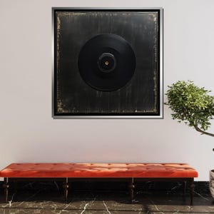 K&ouml;nnte beinhalten: Eine schwarze Schallplatte in einem silbernen Rahmen an einer wei&szlig;en Wand, mit einer roten Bank im Vordergrund.