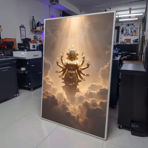 Op de afbeelding: Een ingelijste print met een gouden beeld van een godheid met meerdere armen, tegen een achtergrond van verlichte wolken. Het kunstwerk wordt in een lichte ruimte tentoongesteld, met de nadruk op het hemelse thema en de details.