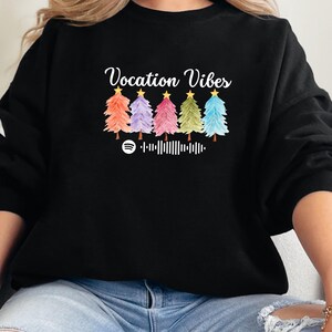 Puede incluir: Sudadera negra con el texto "Vacation Vibes" y una fila de cinco árboles de Navidad coloridos con estrellas en la parte superior. Debajo de los árboles hay un logotipo de Spotify y un código de barras.