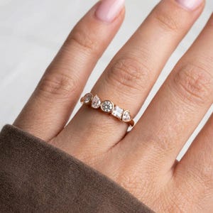 Op de afbeelding: Een gouden ring met een reeks heldere edelstenen in verschillende vormen, waaronder rond, peervormig en hartvormig. De ring wordt getoond op een vinger tegen een neutrale achtergrond.