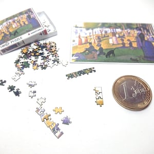 May include: A miniature 180 piece jigsaw puzzle of a painting depicting people gathered in a park. The puzzle pieces are scattered on a white surface next to a small box, a euro coin, and a completed section of the puzzle.