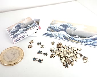 May include: A miniature 160 piece jigsaw puzzle of The Great Wave off Kanagawa by Hokusai, with the box and a few pieces scattered on a white surface. The puzzle is made of wood and has a light brown colour. There is also a Euro coin for scale.