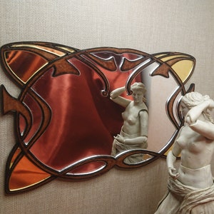 May include: A decorative mirror with a brown wooden frame and intricate carvings. The mirror has a red-tinted glass surface and a gold-colored accent. The mirror is reflecting a statue of a woman.