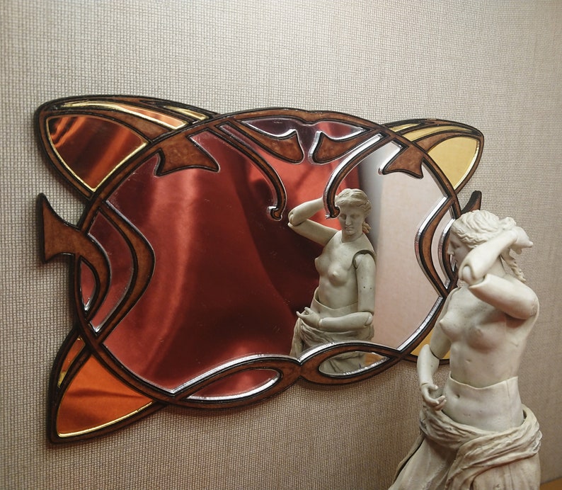 May include: A decorative mirror with a brown wooden frame and intricate carvings. The mirror has a red-tinted glass surface and a gold-colored accent. The mirror is reflecting a statue of a woman.