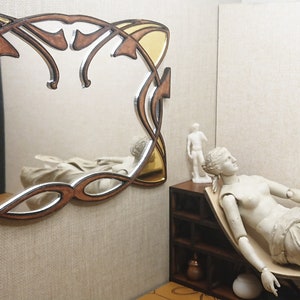 May include: A decorative mirror with a gold and brown wooden frame. The frame has a unique, intricate design with a Celtic knot pattern. The mirror is hanging on a wall with a beige texture.