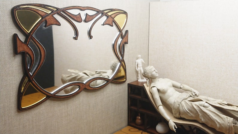 May include: A decorative mirror with a gold and brown wooden frame. The frame has a unique, intricate design with a Celtic knot pattern. The mirror is hanging on a wall with a beige texture.