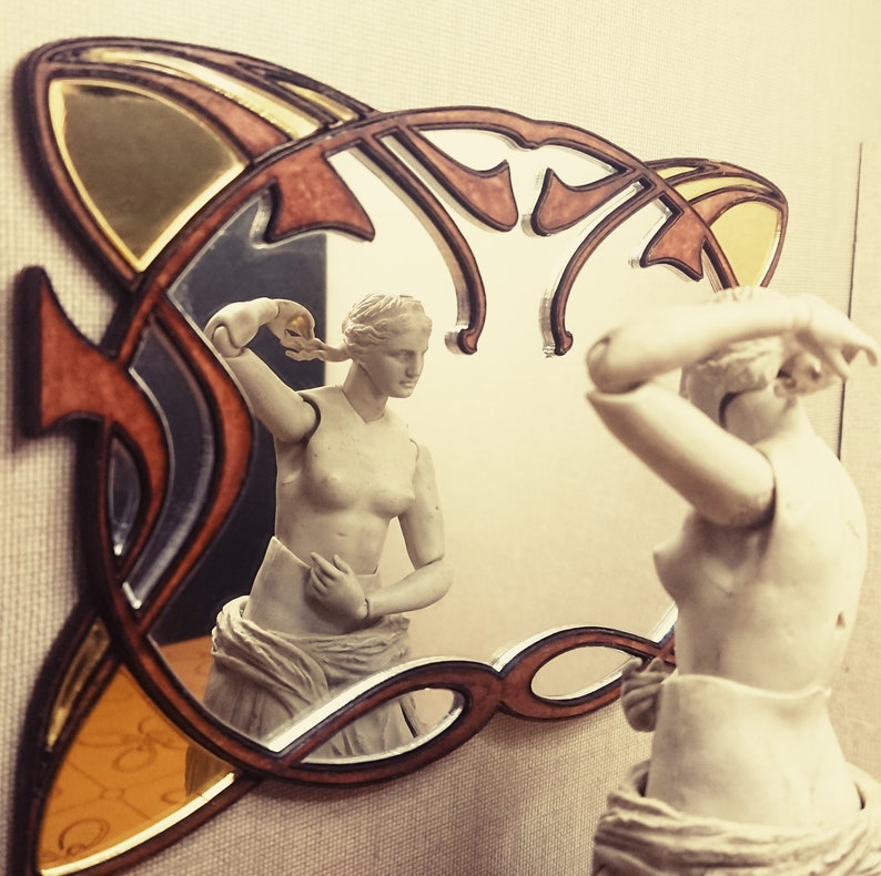 May include: A decorative mirror with a carved wooden frame in the shape of a stylized floral design. The mirror reflects a white statue of a woman with her hair pulled back.