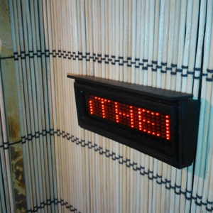 May include: A black rectangular digital display with red LED lights showing the time "11:41". The display is mounted on a wall with a wood slat background.
