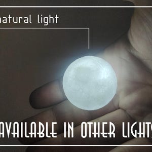 Op de afbeelding: Een gloeiende, bolvormige lichtbron, die lijkt op een kleine maan, in een hand gehouden. De tekst "natural light" en "AVAILABLE IN OTHER LIGHTS" zijn in het wit weergegeven. Het licht straalt zacht wit.