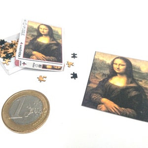 Puede incluir: Una caja miniatura de un rompecabezas de 80 piezas con la pintura de la Mona Lisa. La caja es roja y blanca con las palabras "Amatheria" y "80 Puzzle" impresas en ella. También hay algunas piezas de rompecabezas sueltas esparcidas alrededor de la caja.
