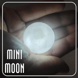 May include: A small, glowing moon-shaped lamp held in a hand. The lamp emits a soft, white light and has a textured surface. The words "MINI MOON" are stacked vertically in a stylized font. The lamp is a decorative item.