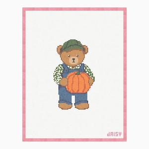 Op de afbeelding: Een kruissteekpatroon op witte stof met een roze rand, met een bruine teddybeer die een groene pet, een groen overhemd met patroon en een blauwe tuinbroek draagt en een grote oranje pompoen vasthoudt. Het woord daisy is onderaan in roze geborduurd.