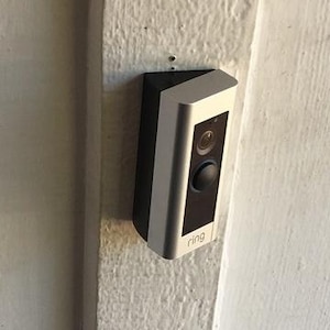 May include: A black and white Ring video doorbell mounted on a textured white wall. The doorbell has a camera lens, a black button, and the brand name "ring" printed on the front.