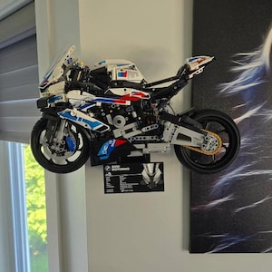 Könnte beinhalten: Ein weißes, blaues und rotes LEGO® BMW Motorrad Modellmotorrad mit schwarzem Rahmen und gelber Kette. Das Motorrad ist an einer weißen Wand montiert.