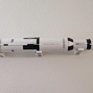Puede incluir: Un modelo Lego blanco y negro del cohete Saturno V, montado en una pared. El cohete tiene una bandera estadounidense roja, blanca y azul en el lateral.