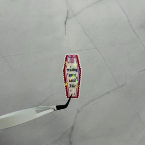 May include: A coffin-shaped sticker with a pink bow at the top and the text "reading smut until 3 die!" in a gothic font. The sticker has a holographic, iridescent finish and a red border. The sticker is being held by a pair of tweezers.