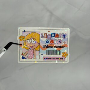 May include: A rectangular holographic sticker designed as a library card. It features a cartoon illustration of a person with yellow hair, the text LIBRARY CARD, LIFETIME MEMBER, and member id: bookbabe, along with a barcode and orange flower icons.