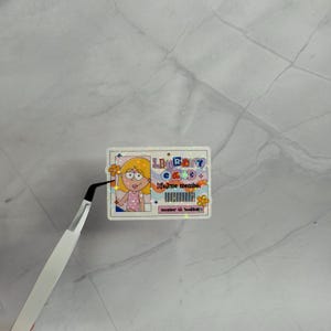 May include: A rectangular holographic sticker designed as a library card with a cartoon illustration of a person with blonde hair, the text "LIBRARY CARD lifetime member", a barcode, and the text "member id: bookbat:)" with orange flower accents.