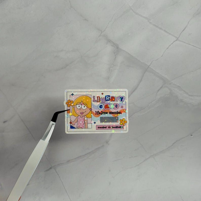 May include: A rectangular holographic sticker designed as a library card with a cartoon illustration of a person with blonde hair, the text "LIBRARY CARD lifetime member", a barcode, and the text "member id: bookbat:)" with orange flower accents.