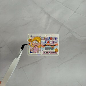 May include: A rectangular holographic sticker designed as a library card with a cartoon illustration of a person with yellow hair, the text "LIBRARY CARD", "lifetime member", and "member id: bookbabe", featuring a barcode and orange flower icons.