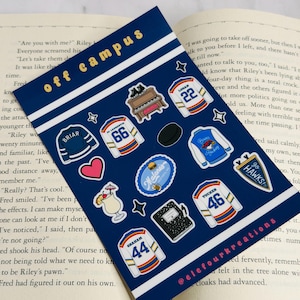 May include: A dark blue sticker sheet featuring the text off campus and elefourkreations. The sheet includes illustrations of hockey jerseys with numbers 22, 66, 46, and 44, a piano with skates, a hockey puck, a heart, a cocktail, a notebook, and a pennant.
