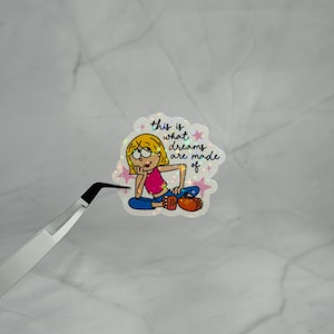 May include: A holographic sticker featuring a cartoon character with blonde hair, a pink top, and blue pants, sitting with a hand on their chin. The text reads, this is what dreams are made of, surrounded by small pink stars on a shimmering background.
