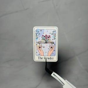 May include: A rectangular sticker featuring a tarot card design. The card depicts a pair of hands holding an open book with flowers blooming from it. The card has a light blue background with stars and the words "The Reader".