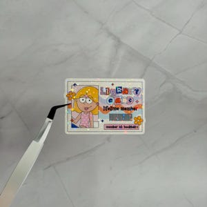 May include: A rectangular holographic sticker designed as a library card. It features a cartoon character with yellow hair, a pink polka dot top, and orange flowers. The text reads LIBRARY CARD, lifetime member, and member id: bookbab.