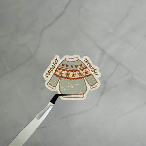 以下が含まれることがあります： ライトグリーンのベースに小さな赤いハートと黄色い花の模様が描かれた、セーター型のホログラムステッカーです。茶色の筆記体で「sweater weather」という文字がプリントされており、表面にはきらめく星のようなエフェクトが施されています。