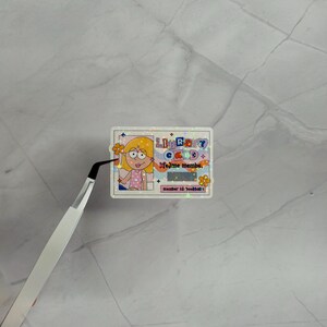 May include: A rectangular holographic sticker designed as a library card with a cartoon illustration of a person with blonde hair. The text reads LIBRARY CARD, lifetime member, and member id: bookbab. It features orange flowers and a barcode.