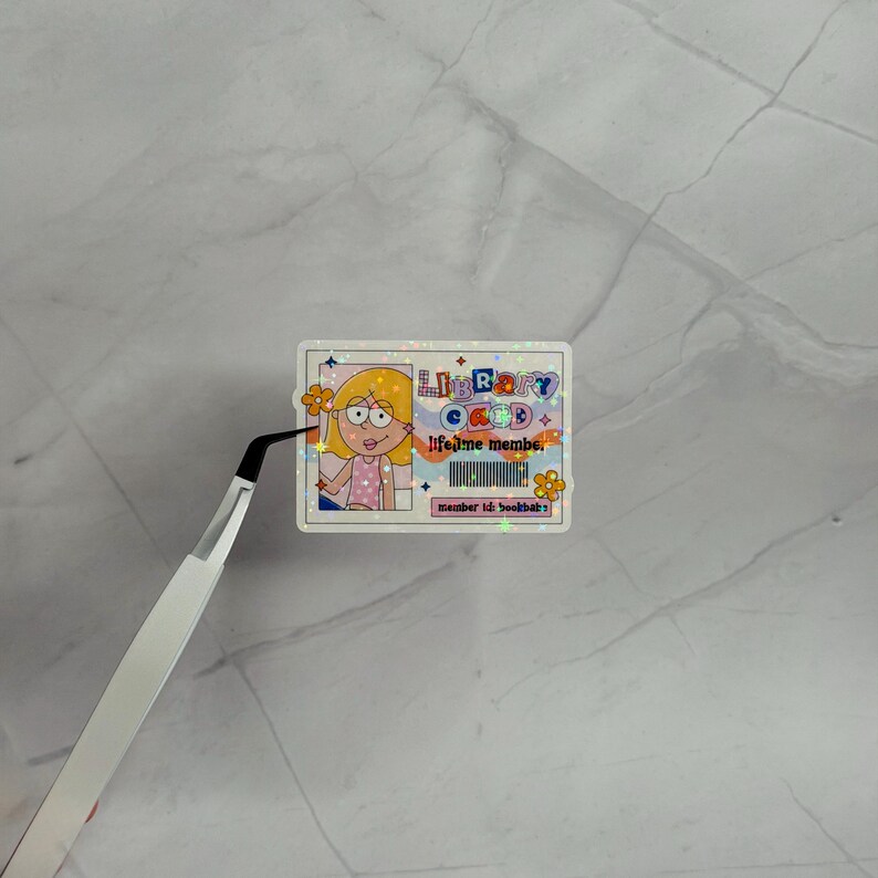May include: A rectangular holographic sticker designed as a library card with a cartoon illustration of a person with blonde hair. The text reads LIBRARY CARD, lifetime member, and member id: bookbab. It features orange flowers and a barcode.