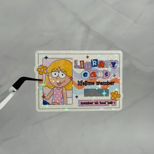 May include: A rectangular holographic sticker designed as a library card featuring a cartoon character with yellow hair and a pink polka dot top. The text reads LIBRARY CARD, lifetime member, and member id: bookbabe, with a barcode graphic.