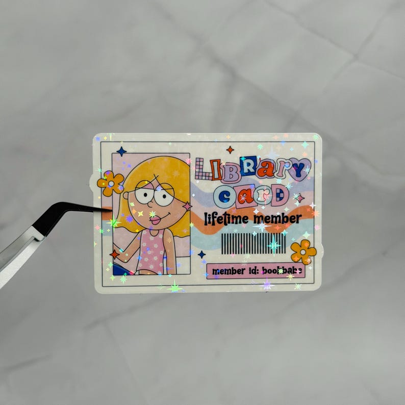 May include: A rectangular holographic sticker designed as a library card featuring a cartoon character with yellow hair and a pink polka dot top. The text reads LIBRARY CARD, lifetime member, and member id: bookbabe, with a barcode graphic.