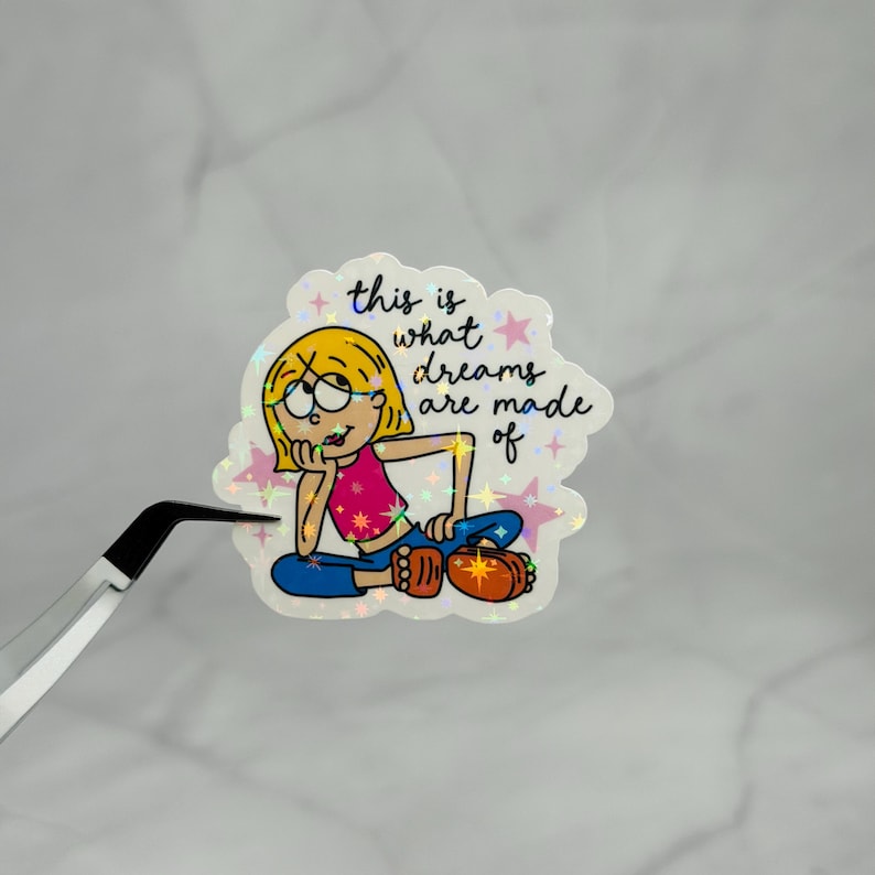 May include: A holographic sticker featuring a cartoon character with blonde hair, a pink top, and blue pants, resting their chin on a hand. The text reads, this is what dreams are made of, surrounded by small pink stars and holographic star-shaped sparkles.