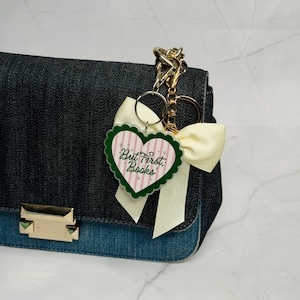 May include: A dark blue denim handbag featuring a gold-toned metal clasp and a decorative keychain. The keychain includes a heart-shaped acrylic charm with pink stripes and the text But First, Books, accented by a cream-colored satin ribbon bow.