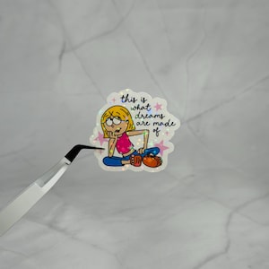 May include: A holographic sticker featuring a cartoon character with blonde hair, a pink top, and blue pants, resting their chin on a hand. The text reads, this is what dreams are made of, surrounded by small pink stars on a white background.