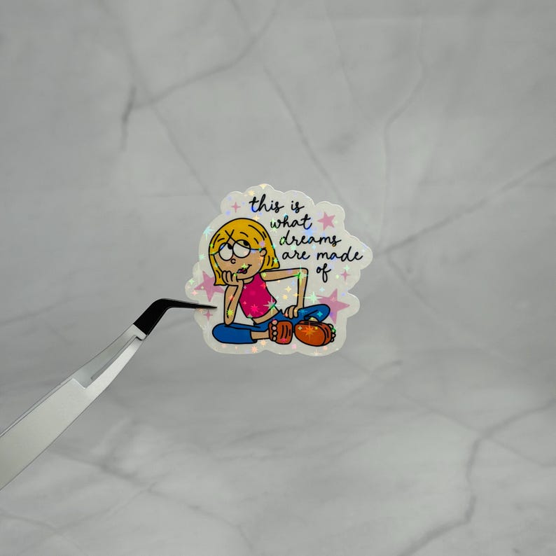 May include: A holographic sticker featuring a cartoon character with blonde hair, a pink top, and blue pants, resting their chin on a hand. The text reads, this is what dreams are made of, surrounded by small pink stars on a white background.